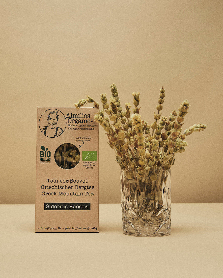 Aimilios Organics griechischer Bergtee auf braunem Hintergrund mit Tee als Dekoration im Glas daneben